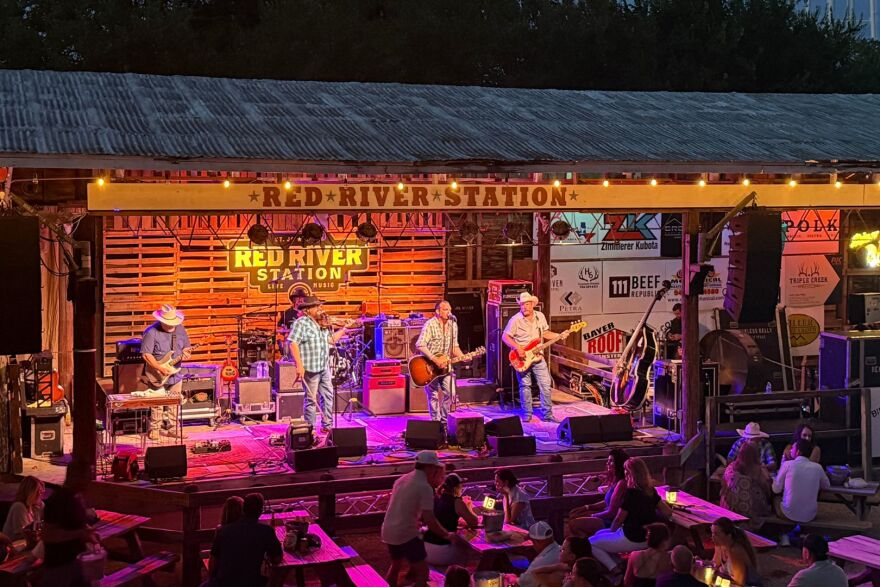 Five-piece band on the Red River Station stage at dusk with sponsor banners along the fence